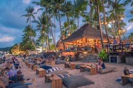 A lively beachside bar filled with people enjoying an evening gathering. There are string lights creating a warm ambiance, palm trees all around, and several large cushions and tables on the sand. The hut-like bar is bustling with activity and decorated with lights, enhancing the tropical atmosphere.