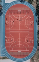 An aerial view of a large, vibrant sports complex featuring a red basketball court with multiple hoops and a surrounding blue running track. Several people are scattered across the area, engaged in various activities.