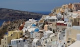 A picturesque hillside town with clusters of whitewashed buildings and blue-domed churches overlooking a deep blue sea. The architecture is characterized by rounded edges and pastel shades, enhancing the scenic beauty of the coastal landscape.