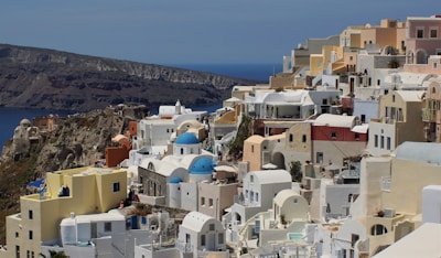 A picturesque hillside town with clusters of whitewashed buildings and blue-domed churches overlooking a deep blue sea. The architecture is characterized by rounded edges and pastel shades, enhancing the scenic beauty of the coastal landscape.