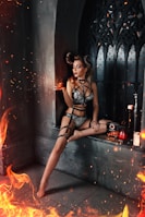 A woman wearing intricate black lingerie and horn accessories poses in a Gothic, stone setting with flames around her. She sits on a ledge next to dark, ornate windows, holding a small orb that emits light and sparks. Candles and a decorative red object with a candle by her side add to the mysterious, fiery atmosphere.