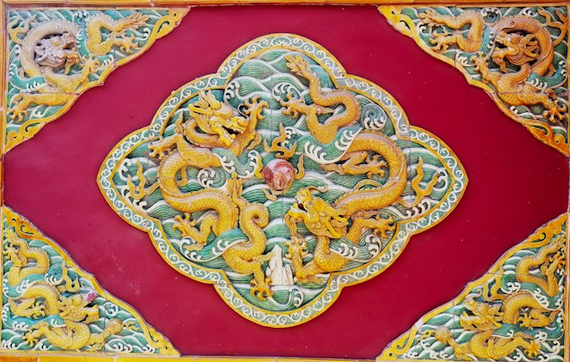 Intricate golden dragons are etched against a deep red background. These dragons are arranged in a symmetrical design, showcasing swirling patterns and detailed scales. The artwork features flowing waves and ornamental borders with fine craftsmanship.