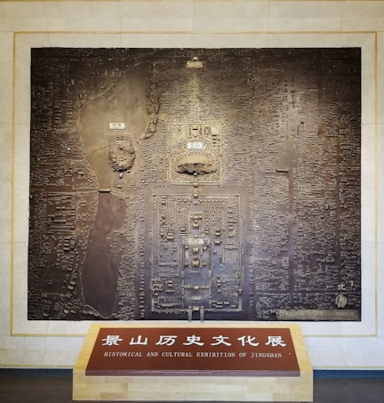 A large, detailed relief map is mounted on a beige wall. The map seems to represent a historical or cultural site, possibly an ancient city or archaeological location, with intricate carvings and structures. Below the map, there is a wooden board with inscriptions in both Chinese and English, stating: 'Historical and Cultural Exhibition of Jingshan.'