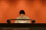 A person wearing a light-colored uniform is standing behind a polished, dark wooden podium against a warm, orange background. The individual appears to be looking down, possibly reading or focusing on something atop the podium.