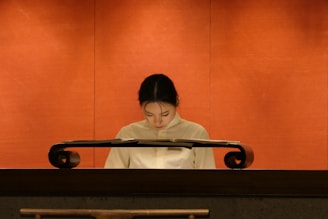 A person wearing a light-colored uniform is standing behind a polished, dark wooden podium against a warm, orange background. The individual appears to be looking down, possibly reading or focusing on something atop the podium.