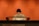 A person wearing a light-colored uniform is standing behind a polished, dark wooden podium against a warm, orange background. The individual appears to be looking down, possibly reading or focusing on something atop the podium.