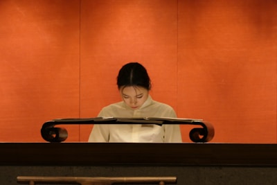 A person wearing a light-colored uniform is standing behind a polished, dark wooden podium against a warm, orange background. The individual appears to be looking down, possibly reading or focusing on something atop the podium.