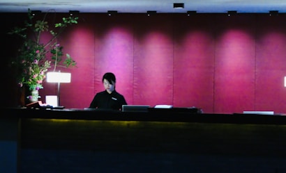 A person is standing behind a dark-colored reception desk, appearing focused and engaged in their task. The background features a deep red wall with an elegant finish, complemented by subtle lighting that creates a warm ambiance. On the desk, there is a lamp and a decorative arrangement of green and pink flowers.