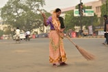 A woman dressed in a traditional orange and green sari is sweeping a street with a large broom. She appears to be focused on her task in an urban setting with light traffic. In the background, there are people on bicycles and motorcycles, as well as a few cars. Trees and commercial buildings with signage are visible, creating a bustling street atmosphere.