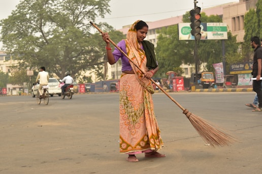 A woman dressed in a traditional orange and green sari is sweeping a street with a large broom. She appears to be focused on her task in an urban setting with light traffic. In the background, there are people on bicycles and motorcycles, as well as a few cars. Trees and commercial buildings with signage are visible, creating a bustling street atmosphere.