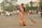 A woman dressed in a traditional orange and green sari is sweeping a street with a large broom. She appears to be focused on her task in an urban setting with light traffic. In the background, there are people on bicycles and motorcycles, as well as a few cars. Trees and commercial buildings with signage are visible, creating a bustling street atmosphere.
