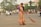 A woman dressed in a traditional orange and green sari is sweeping a street with a large broom. She appears to be focused on her task in an urban setting with light traffic. In the background, there are people on bicycles and motorcycles, as well as a few cars. Trees and commercial buildings with signage are visible, creating a bustling street atmosphere.