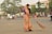 A woman dressed in a traditional orange and green sari is sweeping a street with a large broom. She appears to be focused on her task in an urban setting with light traffic. In the background, there are people on bicycles and motorcycles, as well as a few cars. Trees and commercial buildings with signage are visible, creating a bustling street atmosphere.