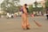 A woman dressed in a traditional orange and green sari is sweeping a street with a large broom. She appears to be focused on her task in an urban setting with light traffic. In the background, there are people on bicycles and motorcycles, as well as a few cars. Trees and commercial buildings with signage are visible, creating a bustling street atmosphere.