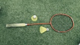 A badminton racket with a red frame and a black grip is lying on a green grass surface. Nearby are two yellow shuttlecocks, one standing upright and the other on its side.