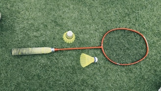 A badminton racket with a red frame and a black grip is lying on a green grass surface. Nearby are two yellow shuttlecocks, one standing upright and the other on its side.