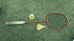 A badminton racket with a red frame and a black grip is lying on a green grass surface. Nearby are two yellow shuttlecocks, one standing upright and the other on its side.