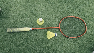 A badminton racket with a red frame and a black grip is lying on a green grass surface. Nearby are two yellow shuttlecocks, one standing upright and the other on its side.