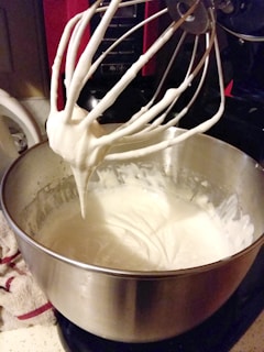 A shiny metal mixing bowl partially filled with a thick, creamy white substance, likely whipped cream or batter. A metal whisk attachment, coated in the same substance, is suspended over the bowl. The scene is set in a kitchen environment with a countertop visible.