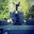 A stone monument in an outdoor setting with a statue of an angel with outstretched wings standing on top of a pedestal. Below the angel, there is a seated figure draped in a cloak, facing the monument. The monument is surrounded by lush green trees in the background.