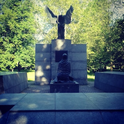 A stone monument in an outdoor setting with a statue of an angel with outstretched wings standing on top of a pedestal. Below the angel, there is a seated figure draped in a cloak, facing the monument. The monument is surrounded by lush green trees in the background.