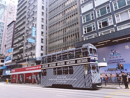 A double-decker tram with modern branding and patterns is traveling along a city street, surrounded by tall buildings. The tram has 'ABCDIOR' written on it in multiple places. Bright advertising banners and signs are visible on the surrounding buildings. People are seen on the sidewalk, and there is a sense of busy urban life.