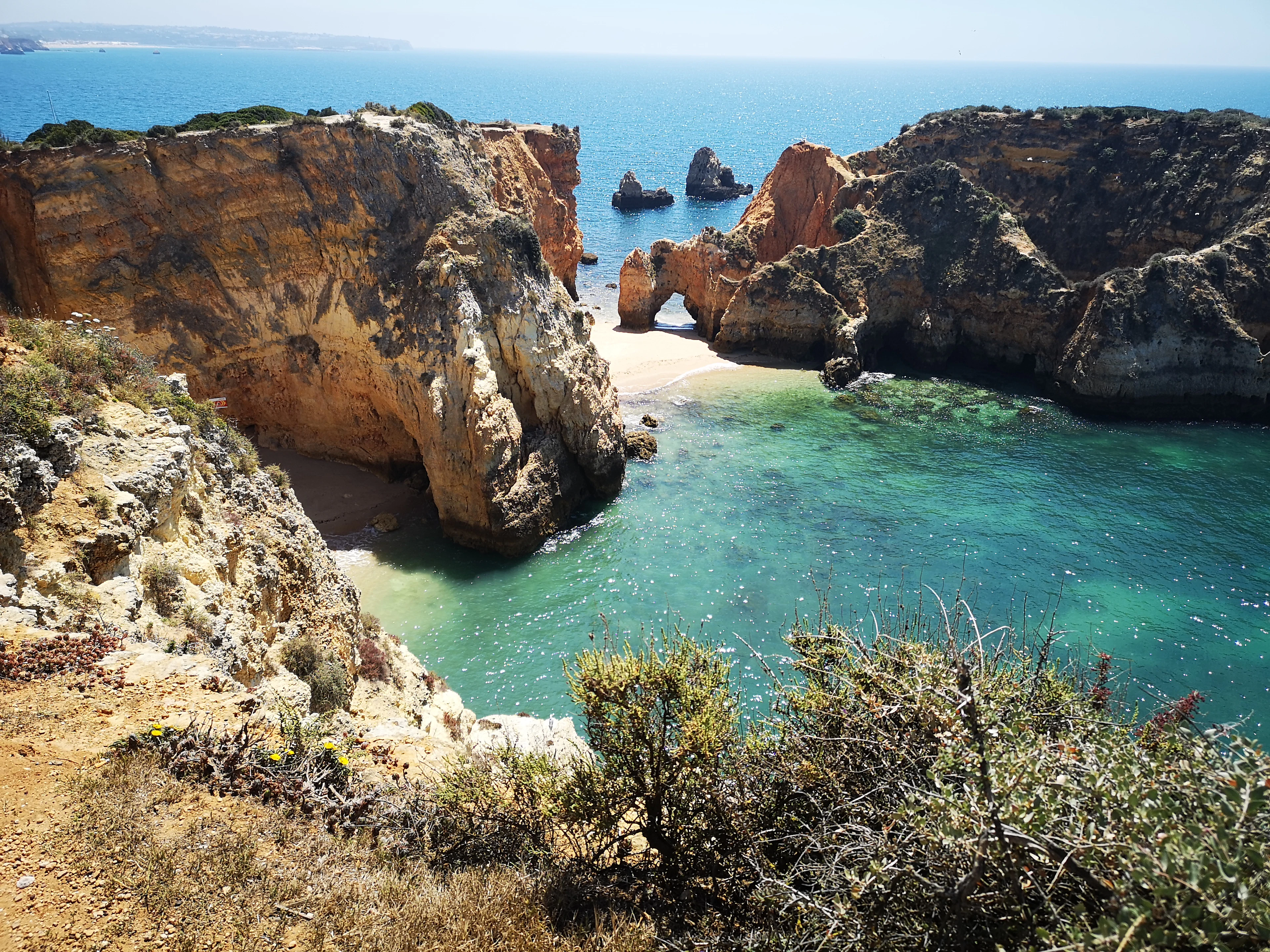 A natural rock arch frames a small sandy cove with turquoise water on the Algarve coast.