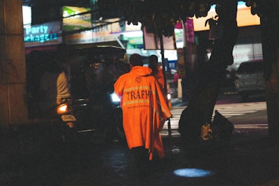 A person wearing an orange traffic safety coat walks along a dimly lit urban street. In the background, blurred figures and vehicles are present, with a noticeable neon-lit signboard illuminating the area.