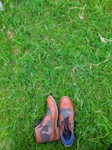 A pair of stylish leather shoes with a mix of brown and camo patterns is placed on lush green grass. The shoes feature intricate detailing and seem to be made for both fashion and durability.