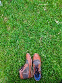 A pair of stylish leather shoes with a mix of brown and camo patterns is placed on lush green grass. The shoes feature intricate detailing and seem to be made for both fashion and durability.