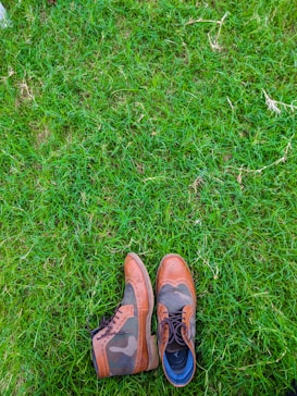 A pair of stylish leather shoes with a mix of brown and camo patterns is placed on lush green grass. The shoes feature intricate detailing and seem to be made for both fashion and durability.