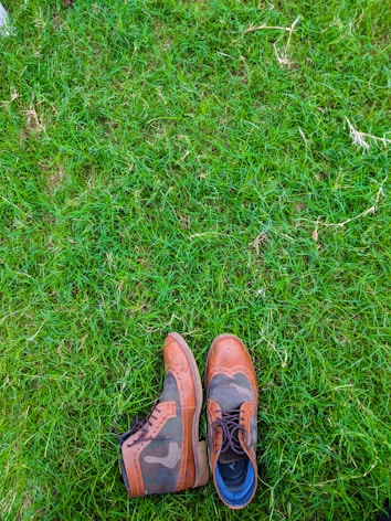 A pair of stylish leather shoes with a mix of brown and camo patterns is placed on lush green grass. The shoes feature intricate detailing and seem to be made for both fashion and durability.