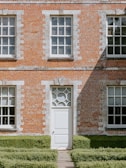 A brick building facade with three large windows and a central door featuring a decorative circular design above it. The lower part of the building is bordered by neatly trimmed green hedges, and the wall shows a mix of orange and white bricks. Sunlight casts sharp shadows across the wall, adding a sense of depth.