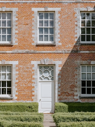 A brick building facade with three large windows and a central door featuring a decorative circular design above it. The lower part of the building is bordered by neatly trimmed green hedges, and the wall shows a mix of orange and white bricks. Sunlight casts sharp shadows across the wall, adding a sense of depth.
