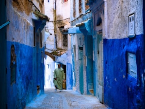 A narrow alleyway with buildings painted in varying shades of blue, characteristic of traditional Moroccan architecture. The pathway has cobblestones, and a person wearing a green robe is walking through the alley. The walls show signs of aging, with some exposed bricks and worn paint.