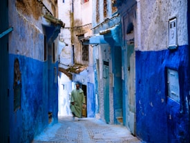 A narrow alleyway with buildings painted in varying shades of blue, characteristic of traditional Moroccan architecture. The pathway has cobblestones, and a person wearing a green robe is walking through the alley. The walls show signs of aging, with some exposed bricks and worn paint.