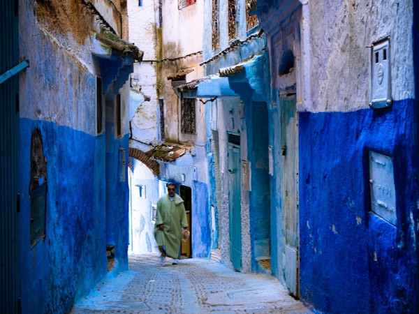 A narrow alleyway with buildings painted in varying shades of blue, characteristic of traditional Moroccan architecture. The pathway has cobblestones, and a person wearing a green robe is walking through the alley. The walls show signs of aging, with some exposed bricks and worn paint.