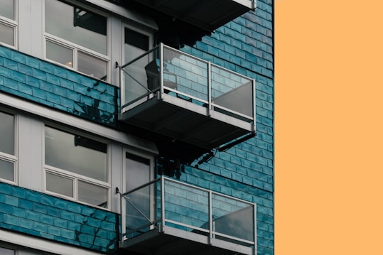 A modern building with reflective glass windows and small metal balconies. The exterior features blue-green tiles, contrasting with an orange background, suggesting a contemporary architectural style.
