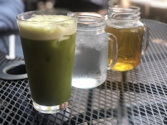 A green matcha latte in a glass is placed on a perforated metal table. Next to it are two mason jars, one containing clear water with ice and the other filled with amber-colored iced tea. The background is slightly blurred, suggesting an outdoor café setting.