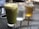 A green matcha latte in a glass is placed on a perforated metal table. Next to it are two mason jars, one containing clear water with ice and the other filled with amber-colored iced tea. The background is slightly blurred, suggesting an outdoor café setting.