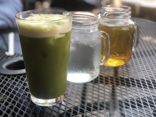 A green matcha latte in a glass is placed on a perforated metal table. Next to it are two mason jars, one containing clear water with ice and the other filled with amber-colored iced tea. The background is slightly blurred, suggesting an outdoor café setting.