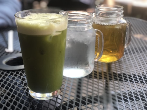 A green matcha latte in a glass is placed on a perforated metal table. Next to it are two mason jars, one containing clear water with ice and the other filled with amber-colored iced tea. The background is slightly blurred, suggesting an outdoor café setting.