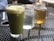 A green matcha latte in a glass is placed on a perforated metal table. Next to it are two mason jars, one containing clear water with ice and the other filled with amber-colored iced tea. The background is slightly blurred, suggesting an outdoor café setting.