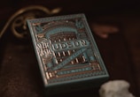 A decorative box of playing cards with intricate, vintage-style designs in gold and blue tones. The word 'Hudson' is prominently displayed on the front along with ornate patterns and architectural images. The background is dark, enhancing the elegance of the packaging.
