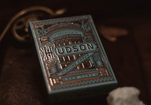 A decorative box of playing cards with intricate, vintage-style designs in gold and blue tones. The word 'Hudson' is prominently displayed on the front along with ornate patterns and architectural images. The background is dark, enhancing the elegance of the packaging.
