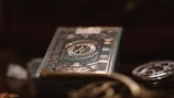 A deck of playing cards with intricate, metallic gold and teal designs featuring geometric patterns and a central emblem. The foreground is slightly out of focus, creating a bokeh effect, with other objects such as coins and a watch faintly visible in the background.