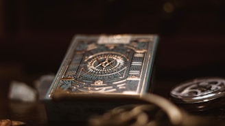 A deck of playing cards with intricate, metallic gold and teal designs featuring geometric patterns and a central emblem. The foreground is slightly out of focus, creating a bokeh effect, with other objects such as coins and a watch faintly visible in the background.