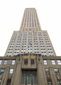 An upward perspective of a tall, iconic skyscraper, emphasizing its towering height and architectural details. The building's facade is adorned with numerous windows arranged in a grid pattern, and the lower part features the words 'EMPIRE STATE' in gold lettering.