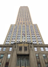 An upward perspective of a tall, iconic skyscraper, emphasizing its towering height and architectural details. The building's facade is adorned with numerous windows arranged in a grid pattern, and the lower part features the words 'EMPIRE STATE' in gold lettering.