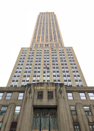 An upward perspective of a tall, iconic skyscraper, emphasizing its towering height and architectural details. The building's facade is adorned with numerous windows arranged in a grid pattern, and the lower part features the words 'EMPIRE STATE' in gold lettering.